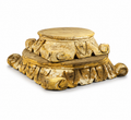 An ornate carved wooden pedestal or display stand with a tiered, acanthus leaf-style base and a round top platform, finished in a distressed gold and brown tone, shown on a white background.